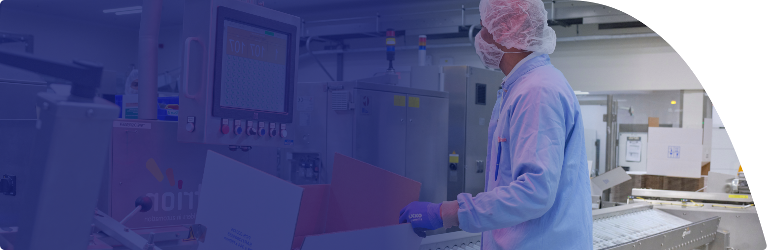 Ein Mitarbeiter in hygienischer Schutzkleidung steht an einer hochmodernen Produktionslinie und überprüft eine Verpackungseinheit. Die Umgebung zeigt automatisierte Geräte und Monitore zur Qualitätskontrolle. 