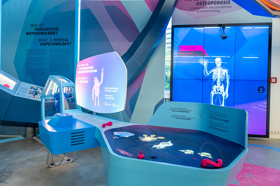 Das Foto zeigt Amgens interaktive Themeninsel zur medizinischen Biotechnologie im Deutschen Museum. Zu sehen sind blaue und pinkfarbene Infotafeln, ein Tisch mit grafischen Darstellungen und ein großer Bildschirm mit einem menschlichen Skelett.