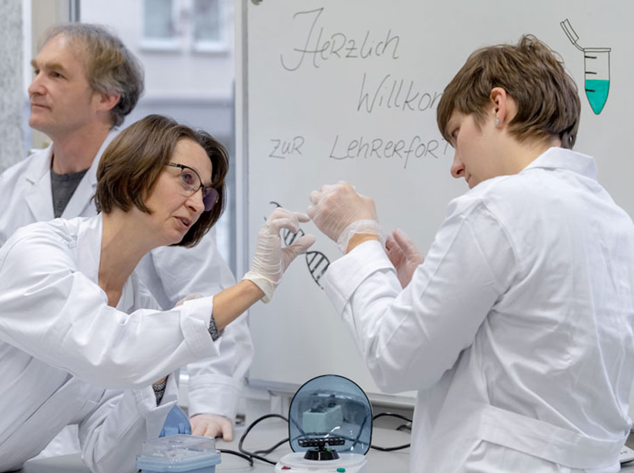 Zwei Personen im weißen Laborkittel im Vordergrund führen ein biotechnologisches Experiment im Labor durch. Im Hintergrund zeigt eine Tafel den Schriftzug ‚Herzlich Willkommen zur Lehrerfortbildung‘, neben der Tafel steht ein Mann. 
