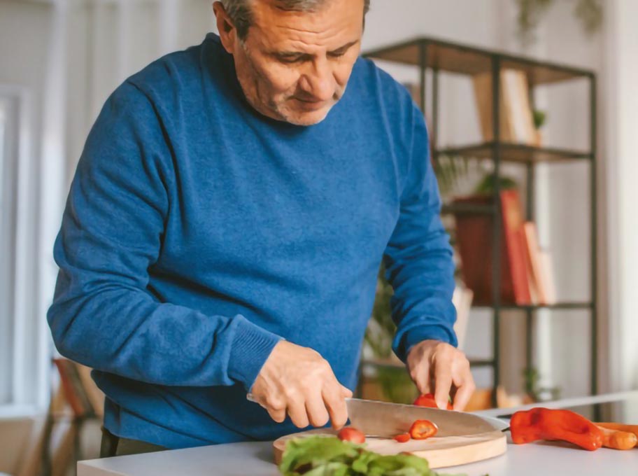 Ein älterer Mann steht an einer Kücheninsel und schneidet eine rote Paprika auf einem Holzbrett. Im Hintergrund ist verschwommen ein Wohnbereich zu sehen.