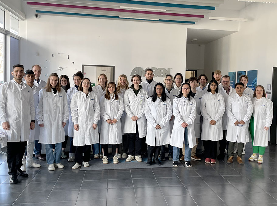 Ein Gruppenfoto von ca. 25 Personen in Kitteln im Eingangsbereich des Amgen Forschungsstandorts in München. Man sieht Stipendiat:innen und Mitarbeitende der Amgen GmbH und der Amgen Research (Munich) GmbH.