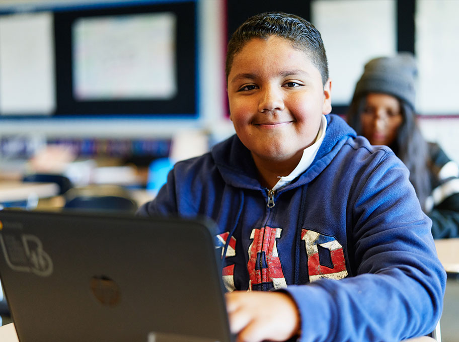 Ein Junge sitzt in einem Klassenzimmer an einem Laptop und lächelt in die Kamera. Hinter ihm sind verschwommen ein Mädchen, mehrere Schulbänke und Plakate zu sehen.